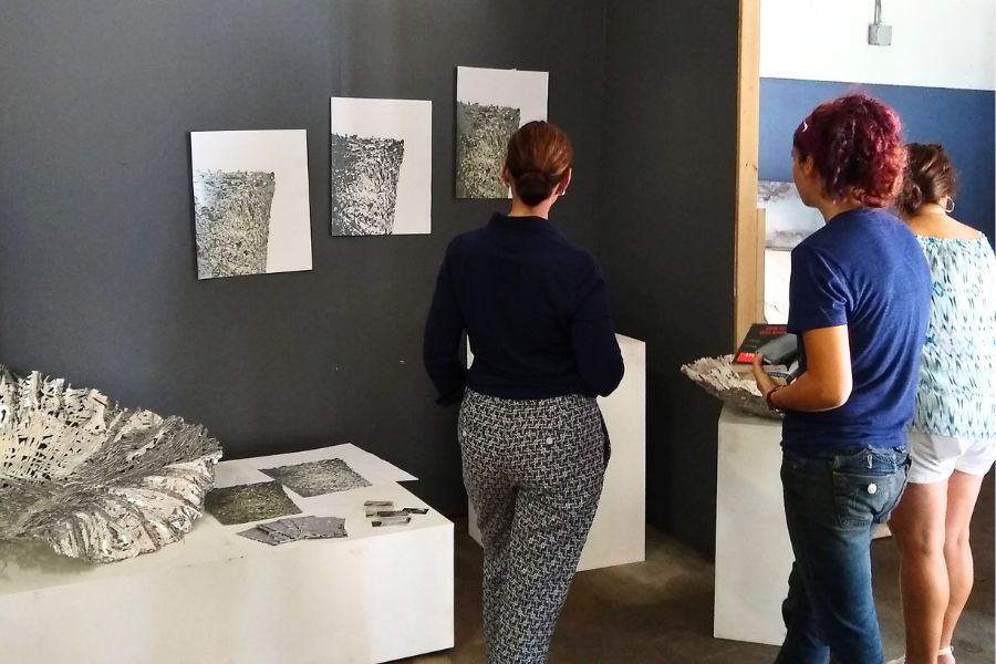exhibit A few people standing looking at a gallery wall withe three pieces of art work and a large metal sculpture.